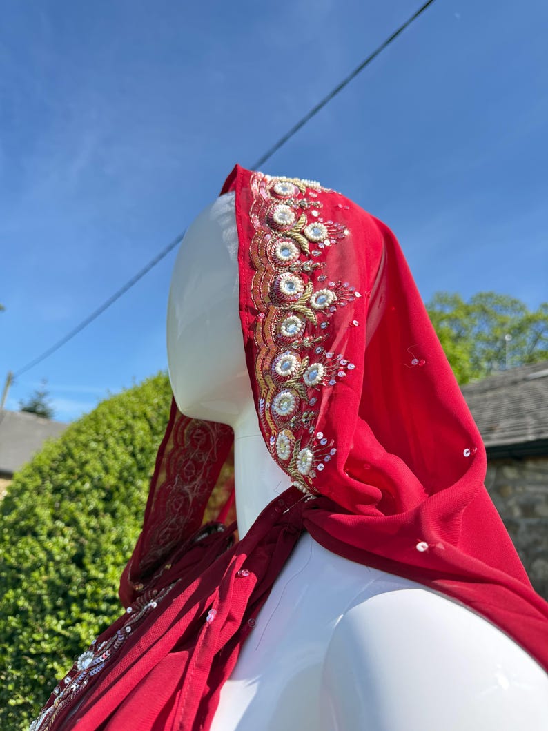 May include: A red sheer cape with gold and silver embroidery. The cape has a hood and is decorated with sequins and beads.