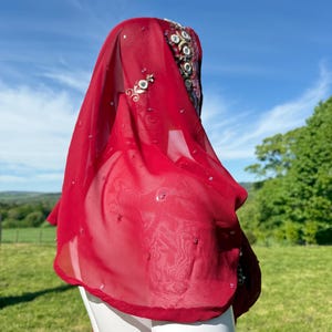 May include: A red sheer scarf with beaded embellishments. The scarf is draped over a white mannequin's head and shoulders.