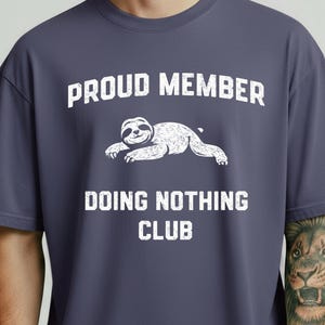 Op de afbeelding: Denim blauw T-shirt met de witte tekst "PROUD MEMBER DOING NOTHING CLUB". Een cartoon luiaard is afgedrukt boven de tekst "DOING NOTHING CLUB". Het shirt heeft een relaxte pasvorm.