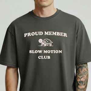 Puede incluir: Camiseta gris oscuro con el texto blanco "PROUD MEMBER SLOW MOTION CLUB". Una tortuga de dibujos animados está encima de las palabras "SLOW MOTION CLUB". La camiseta es de corte holgado.
