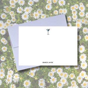 Può includere: Set di cancelleria bianco con un'illustrazione di bicchiere da cocktail e il testo "BIANCA JAYDE". Il set include un biglietto bianco e una busta lilla chiaro. Lo sfondo presenta margherite bianche.