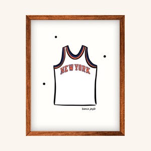 May include: A framed print featuring a white basketball jersey with the text "NEW YORK" in orange and blue. The jersey design is outlined in black. The print is in a brown wooden frame, with the artist's signature at the bottom.