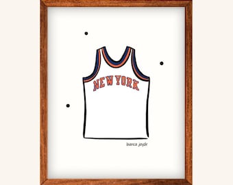 Pode incluir: Uma impressão emoldurada de uma camisa de basquete branca com o texto "NEW YORK" em laranja e azul. O design da camisa é delineado em preto. A impressão está em uma moldura de madeira marrom, com a assinatura do artista na parte inferior.