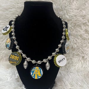May include: A silver chain necklace with various bottle caps attached. The bottle caps feature different designs, including a football, a logo with the letters "AT", and the text "1891 University".