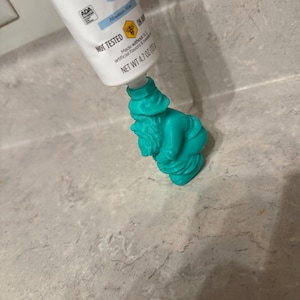 Può includere: Un tubetto di dentifricio Mountain Mint viene spremuto in uno spremi-tubo a forma di gnomo turchese. Il tubetto di dentifricio è bianco con dettagli blu e la scritta "ADA" e "NOT TESTED". Lo spremi-tubo è un dettagliato gnomo da giardino in miniatura.