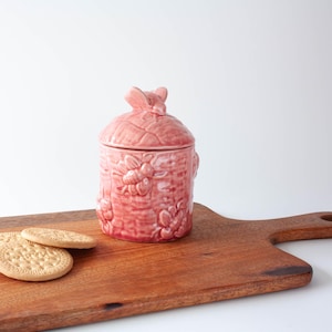 Può includere: Un barattolo di ceramica rosa con coperchio, con un design a forma di ape e favo. Il barattolo è appoggiato su un tagliere di legno, con due biscotti rotondi in primo piano. Il barattolo ha una maniglia a forma di ape sul coperchio.
