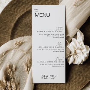 Może przedstawiać: Biała karta menu z czarnym tekstem, prezentująca menu weselne. Menu zawiera przystawki, danie główne i opcje deserów. Karta umieszczona jest na drewnianej ładowarce z lnianą serwetką i ozdobnymi gałązkami.