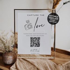 May include: A white sign with black text and a black and white illustration of a camera. The text reads "Capture the love" and "Please share your photos with us! Scan the code and upload." A black and white QR code is below the text. The text "With Thanks" and "Claire & Paul" is at the bottom of the sign. The sign is in a silver frame.