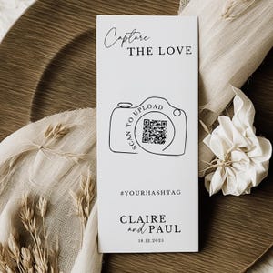 May include: A white wedding invitation card featuring the text "Capture the Love" and a camera graphic with a QR code. The card also includes the text "Scan to Upload", a hashtag, and the names "Claire and Paul" with the date 18.12.2025.