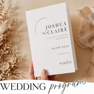 以下が含まれることがあります： 黒い文字で「Joshua and Claire」と日付「05.06.202X」が記載された白いウェディングプログラム。プログラムは手で持たれ、背景には乾燥した植物とベージュの布があります。「WEDDING program」の文字が下にあります。