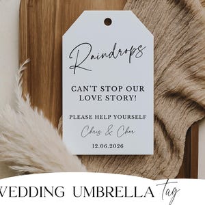 May include: A white tag with the text "Raindrops CAN'T STOP OUR LOVE STORY! PLEASE HELP YOURSELF Chris & Char 12.06.2026". The tag is on a wooden surface with a beige cloth and decorative white feathers. The words "WEDDING UMBRELLA Tag" are at the bottom.