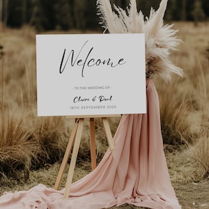 May include: A white wedding welcome sign on a wooden easel, with the word "Welcome" in script. The sign reads "- TO THE WEDDING OF - Claire & Paul 18TH SEPTEMBER 2025". The sign is decorated with pampas grass and draped with a pink fabric.