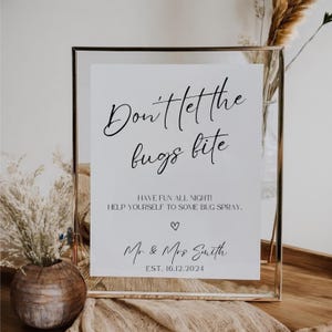 May include: A white sign with black text that reads "Don't let the bugs bite. Have fun all night! Help yourself to some bug spray. ♡ Mr & Mrs Smith EST. 16.12.2024". The sign is framed in a gold frame and is sitting on a table with a brown vase and dried grass.