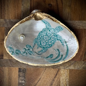May include: A seashell dish with a gold rim, featuring a green and white turtle design with waves and glitter. A white pearl sits inside the shell.