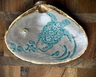 May include: A seashell dish with a gold rim, featuring a green and white turtle design with waves and glitter. A white pearl sits inside the shell.