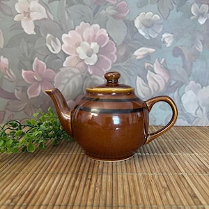 May include: A glossy brown ceramic teapot featuring a rounded body with two horizontal dark brown stripes encircling the upper portion, a curved spout, a matching lid with a small knob, and a sturdy handle on the side for easy pouring.