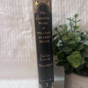 Puede incluir: Un lomo de libro beige con un panel rectangular negro que presenta letras doradas que dicen The Complete Works of WILLIAM SHAKESPEARE, Julius Caesar, Macbeth y VOLUME 2, enmarcado por filigrana dorada decorativa arriba y abajo.