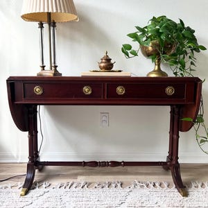 Op de afbeelding: Een mahoniehouten console met twee lades en messing hardware. Een lamp met geplooide lampenkap, een kleine decoratieve pot en een goudkleurige plantenbak met groen staan erop. De tafel heeft klepbladen en staat op een patroonkleed.