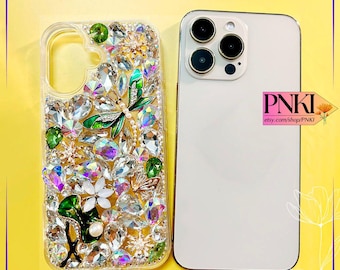May include: Clear phone case bedazzled with iridescent rhinestones, green gems, and a dragonfly design. The case is next to a white smartphone. The text "PNKI" and "Bling Case Diy" are visible.