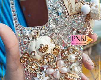 May include: A clear phone case heavily embellished with rhinestones, pearls, and gold accents. Features a white pumpkin carriage, a wooden heart with "I love you" text, and a miniature Eiffel Tower charm. The case is encrusted with sparkling crystals.
