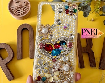 May include: Clear phone case embellished with pearls, rhinestones, and floral accents. A heart-shaped design is centered, filled with red, blue, and clear gems. The case has the text "PNKI" and "Etsy.com/shop/PNKI".