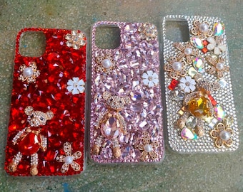 May include: Three bedazzled phone cases. One is red with a teddy bear and floral accents. The second is pink with a teddy bear and floral accents. The third is silver with floral accents.