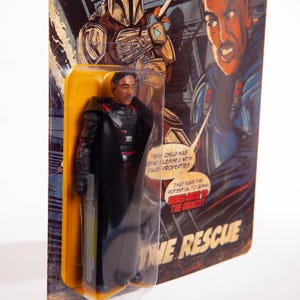 May include: A retro action figure of a character in a black outfit with a long coat, holding a green lightsaber. The figure is in a clear plastic package with a yellow backing card featuring comic book-style art and the text "THE RESCUE".
