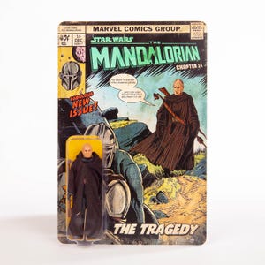 May include: A Star Wars action figure of a character from The Mandalorian, in a clear plastic case, is displayed in front of a vintage comic book cover. The comic book cover features the title "The Mandalorian Chapter 14: The Tragedy."