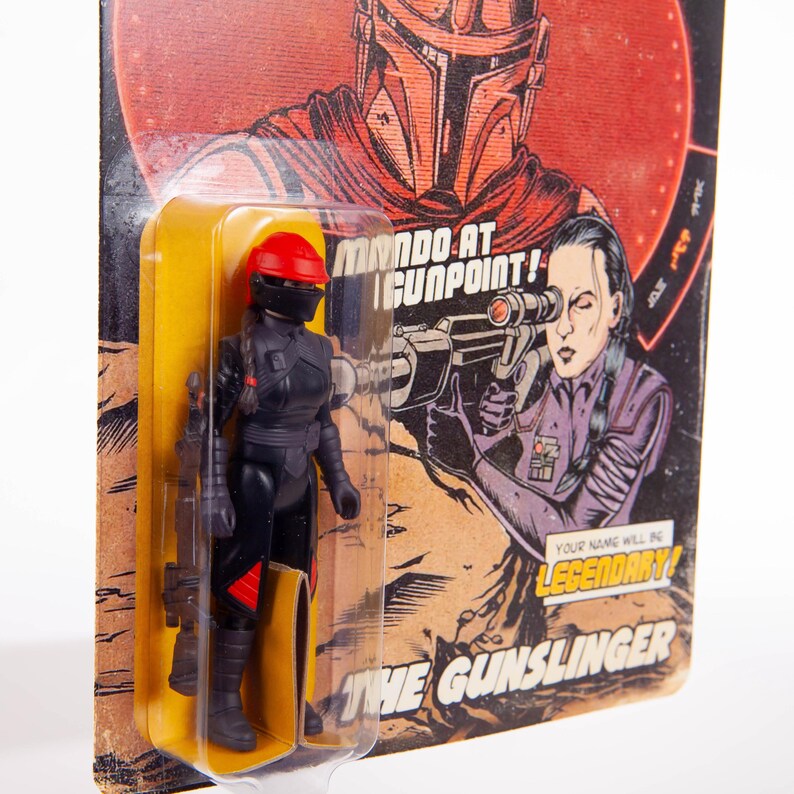 May include: A black action figure of a female character in a red helmet and black uniform, packaged on a vintage-style card. The card features illustrations of a helmeted figure and a woman aiming a weapon, with the text "MANDO AT GUNPOINT!" and "THE GUNSLINGER".