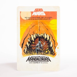 May include: A Star Wars trading card featuring a scene from The Mandalorian. The card has an orange and yellow background with the text "THEY SAY IT LIVES IN THERE. THEY SAY IT SLEEPS. IT LIVES IN AN ABANDONED SARLACC PIT." The Mandalorian is in the foreground.
