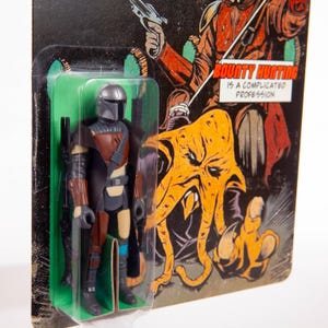 May include: Action figure of a Mandalorian character in a plastic blister pack. The figure is wearing a helmet, armor, and a cape. The packaging features the text "Bounty Hunting Is A Complicated Profession" and an illustration of a creature.