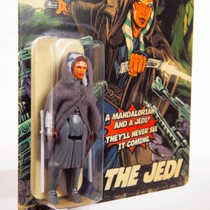 Puede incluir: Figura de acci&oacute;n vintage de un personaje con piel naranja, marcas blancas y una capa gris con capucha. La figura est&aacute; en un paquete de pl&aacute;stico transparente con un fondo de estilo c&oacute;mic. El texto en el embalaje dice: "&iquest;Un Mandaloriano y un Jedi? Nunca lo ver&aacute;n venir."