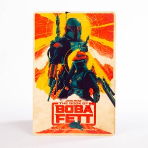May include: A rectangular card featuring Boba Fett and another character from the Star Wars series. The card has a vintage aesthetic with a red and yellow background. The text "STAR WARS THE BOOK OF BOBA FETT" is displayed in red and white.