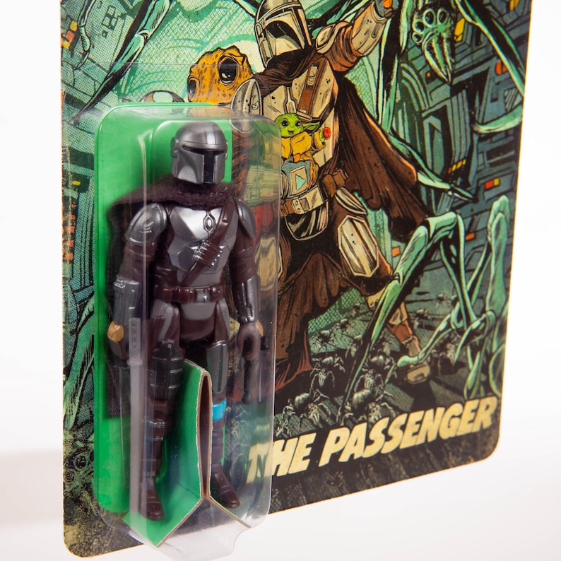 May include: A Mandalorian action figure in a clear plastic package. The figure is dark gray and brown, with a helmet and armor. The packaging features a green and brown illustration of the Mandalorian and the text "THE PASSENGER".