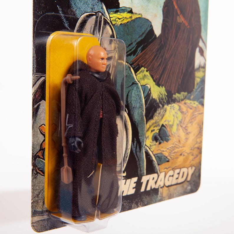 May include: A vintage action figure of a bald character in a dark brown robe, packaged in a yellow and clear plastic blister pack. The card backing features a comic book style illustration with the words "THE TRAGEDY".