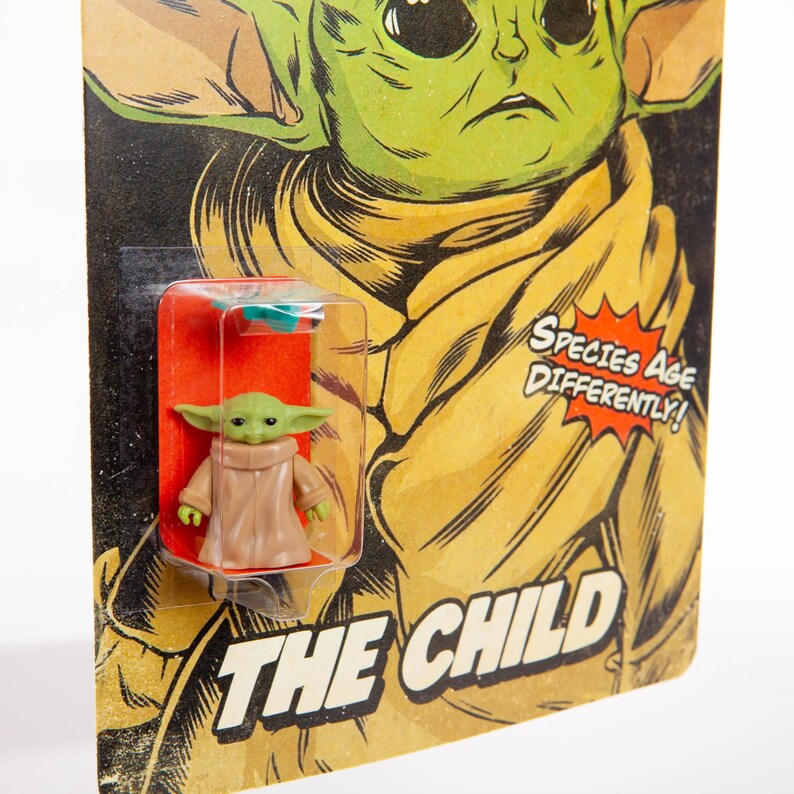 May include: A small green figure of The Child, also known as Baby Yoda, in a clear plastic case on a retro-style card. The card has a yellow and brown background with the words "THE CHILD" and "SPECIES AGE DIFFERENTLY!"