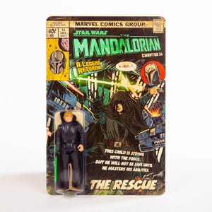 May include: A Star Wars Mandalorian action figure on a vintage-style comic book card. The card features comic book art with the text "The Mandalorian Chapter 16" and "The Rescue". The figure is of a character in a black outfit with a green lightsaber.