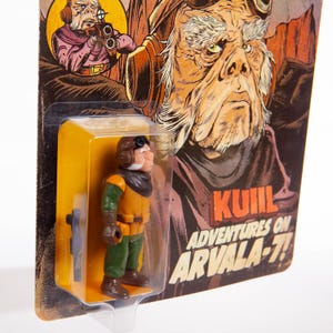 May include: A vintage action figure of a character in a yellow and green uniform, with a brown helmet and a blaster. The figure is packaged on a card with the text "Kuiil Adventures on Arvala-7!" and a character illustration.