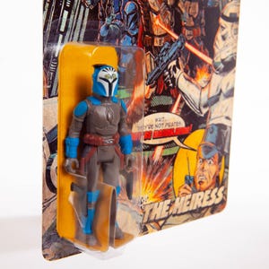 May include: A vintage-style action figure of a character in a blue and gray suit with a helmet. The figure is in a clear plastic blister pack with a yellow backing card. The card features comic book-style artwork and the text "THE HEIRESS."