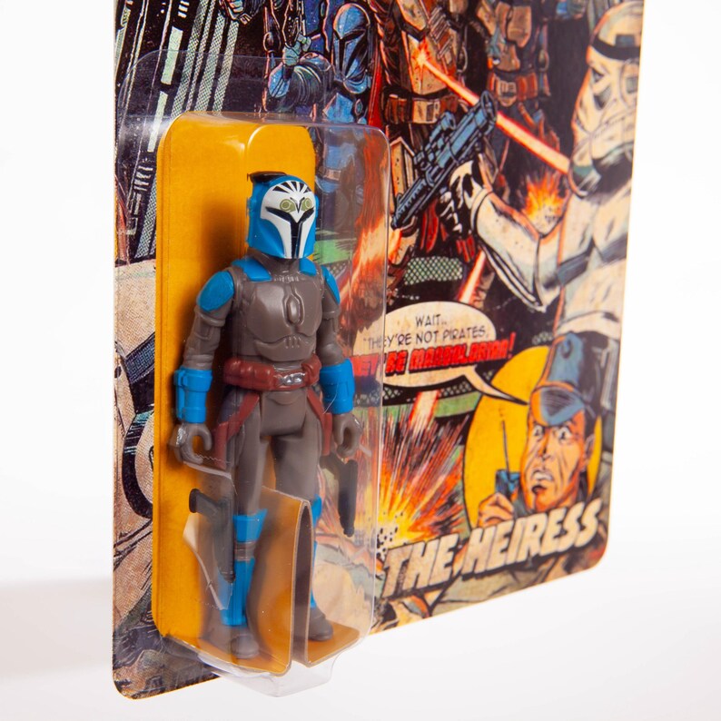 May include: A vintage-style action figure of a character in a blue and gray suit with a helmet. The figure is in a clear plastic blister pack with a yellow backing card. The card features comic book-style artwork and the text "THE HEIRESS."