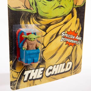 Puede incluir: Una peque&ntilde;a figura verde de The Child, tambi&eacute;n conocido como Grogu, en un portabeb&eacute;s azul y rojo. La figura est&aacute; empaquetada en una tarjeta de estilo vintage con las palabras "THE CHILD" y "Species Age Differently!"