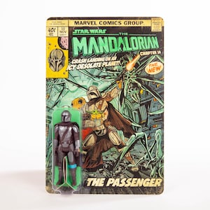 May include: A vintage Star Wars Mandalorian action figure on a comic book-style card. The card features comic book art with the text "The Mandalorian" and "The Passenger." The figure is in a clear plastic blister pack.