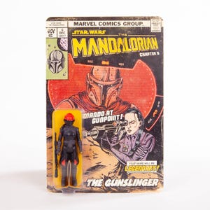May include: A vintage-style Star Wars: The Mandalorian comic book cover with a matching action figure. The comic features a red and black illustration of the Mandalorian and the text "Mando at Gunpoint!" The action figure is black and red.