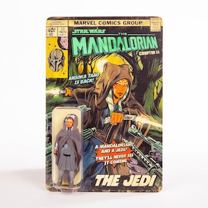 Puede incluir: Una figura de acci&oacute;n vintage de Star Wars de Ahsoka Tano, empaquetada en una tarjeta de respaldo estilo c&oacute;mic. La tarjeta presenta ilustraciones de la serie "The Mandalorian", con el texto "Ahsoka Tano is Back!" y "The Jedi."