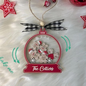 May include: A globe-shaped Christmas ornament with a red frame, featuring the text 2026 and The Colliers. Inside are loose wooden gingerbread figures, paw prints, and hearts. It includes a black and white checkered bow and a jute twine hanger.