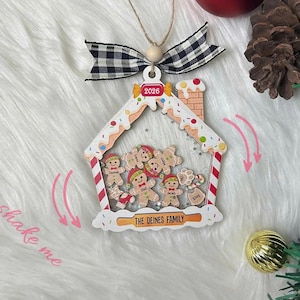 May include: A gingerbread house-shaped Christmas ornament with a clear centre containing small gingerbread person figures. The frame features a black and white buffalo plaid bow, the year 2026, and the text THE DEINES FAMILY and shake me.