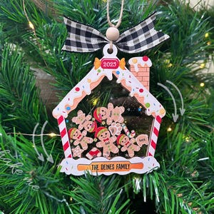 May include: A wooden gingerbread house Christmas ornament with a clear plastic window showing gingerbread people with names. The ornament is decorated with white frosting, red candy canes, and colorful sprinkles. The year 2025 is written on the roof. The text "The Deines Family" is written on the bottom of the ornament.