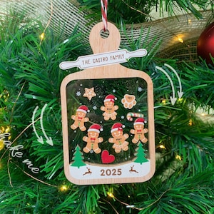 May include: A wooden Christmas ornament with a clear window, featuring gingerbread people, stars, and a heart. The ornament is personalized with "The Castro Family" and the year 2025. It hangs on a Christmas tree with green needles and red and gold ornaments.