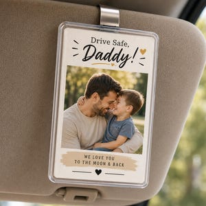 May include: A rectangular car visor clip featuring a photograph of a father and child. The clip is inscribed with the words "Drive Safe, Daddy!" and "We love you to the moon & back". The photo shows a man and a young boy embracing.