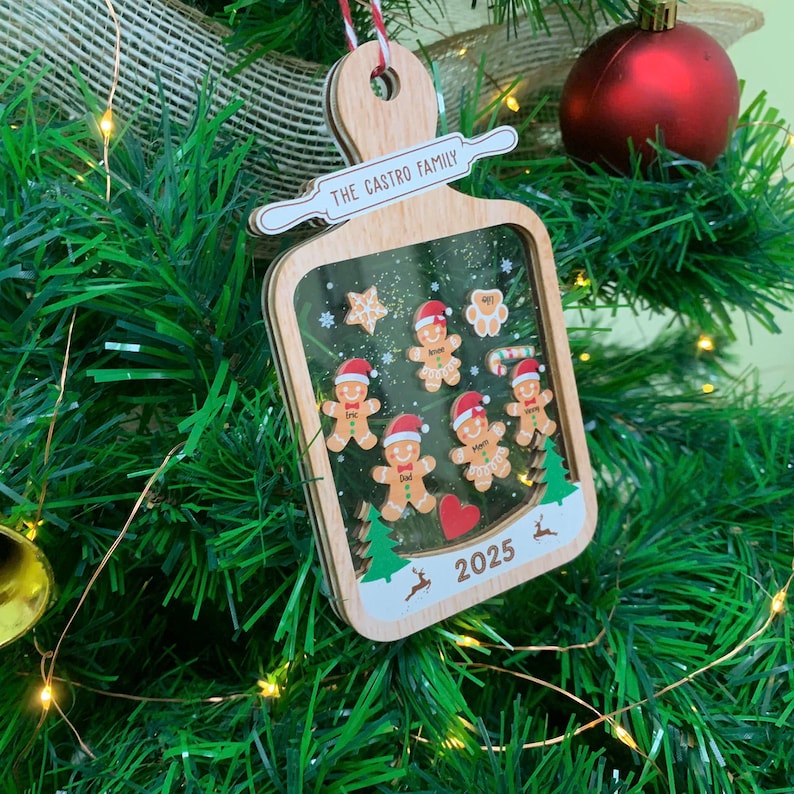 May include: Wooden Christmas ornament with a clear window, featuring gingerbread people, stars, and trees. The ornament has the text "THE CASTRO FAMILY" and "2025". It is hanging on a Christmas tree with a red ornament.